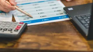 Mão aponta caneta para tabela de taxas ao lado de calculadora e notebook, ilustrando a dedução de JCP