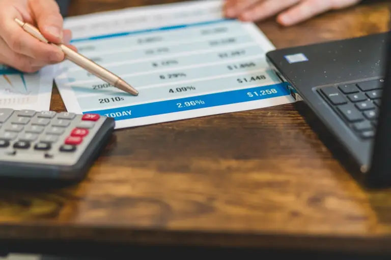 Mão aponta caneta para tabela de taxas ao lado de calculadora e notebook, ilustrando a dedução de JCP